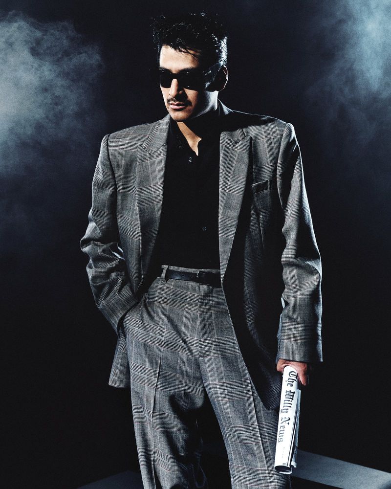 Man wearing a grey plaid suit holding a newspaper against a smoky dark background