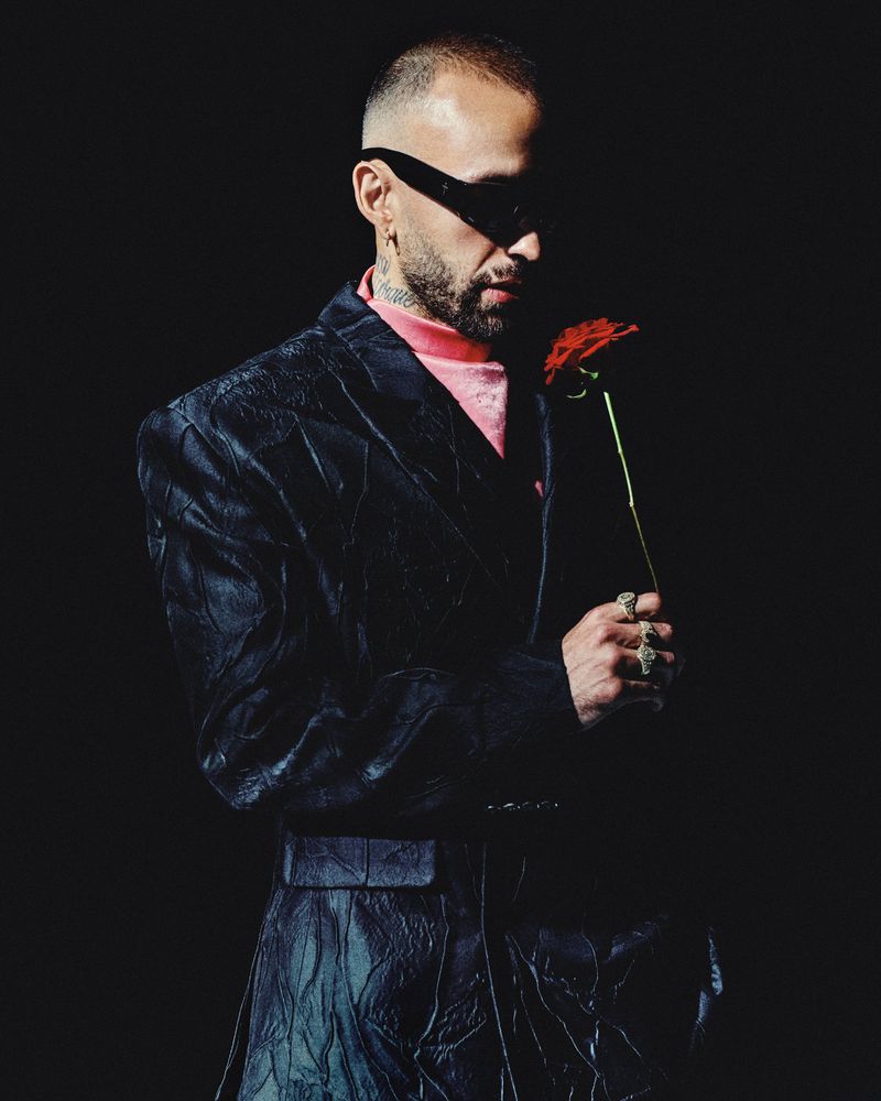 Man wearing dark sunglasses and textured black suit holding a red rose against a black background