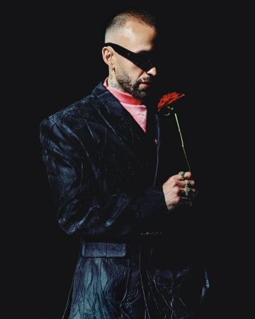 Man wearing dark sunglasses and textured black suit holding a red rose against a black background
