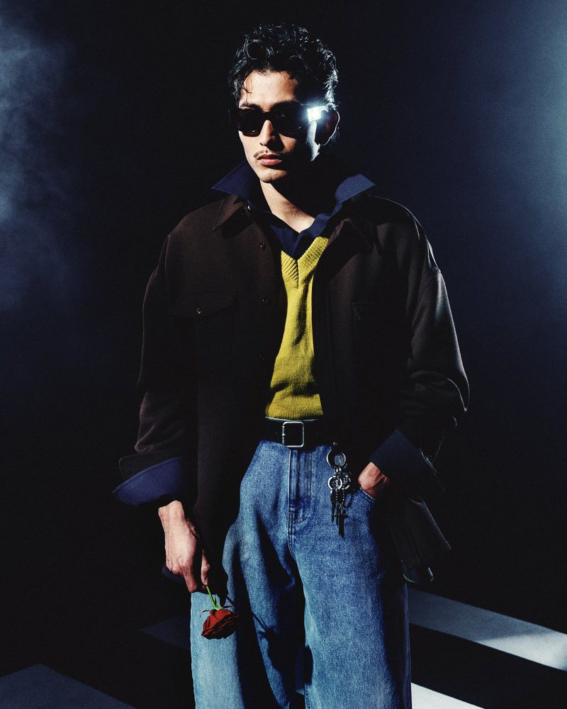 Man wearing dark sunglasses, brown jacket, yellow sweater, and blue jeans holding a red rose against a dark moody background.