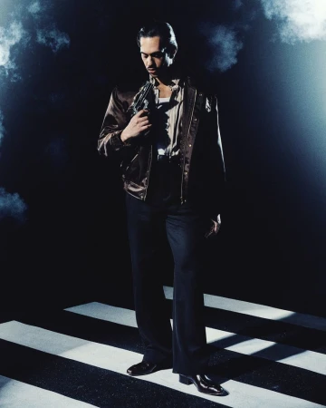 Man wearing a brown satin jacket and dark trousers standing against a dark background with atmospheric smoke