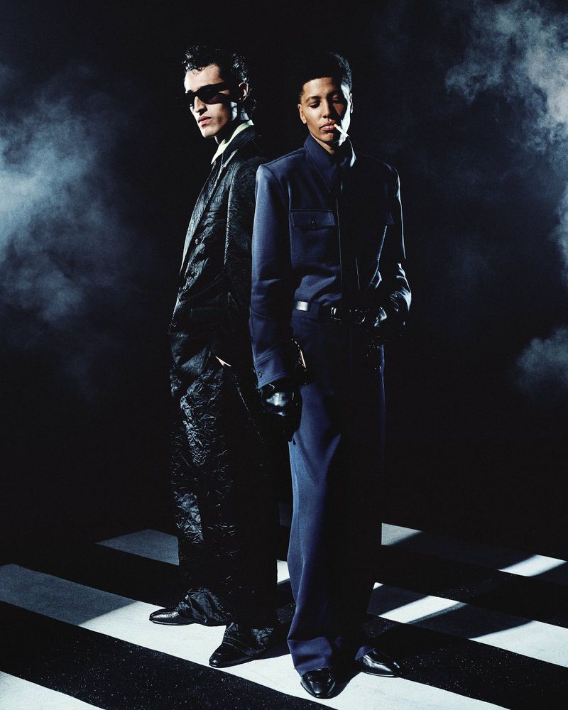 Two models standing back-to-back in dark tailored suits against a smoky, high-contrast studio background