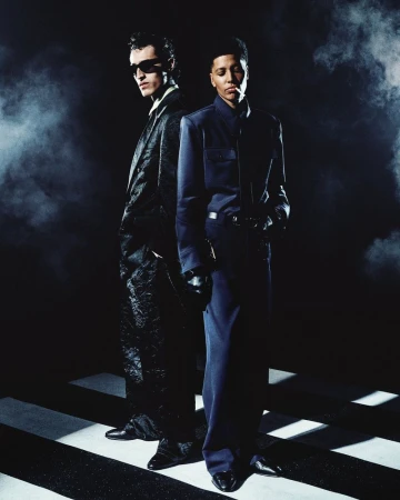 Two models standing back-to-back in dark tailored suits against a smoky, high-contrast studio background