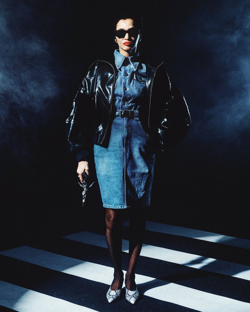 Model wearing dark sunglasses, shiny black leather jacket, and denim outfit standing on striped floor
