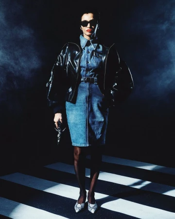 Model wearing dark sunglasses, shiny black leather jacket, and denim outfit standing on striped floor
