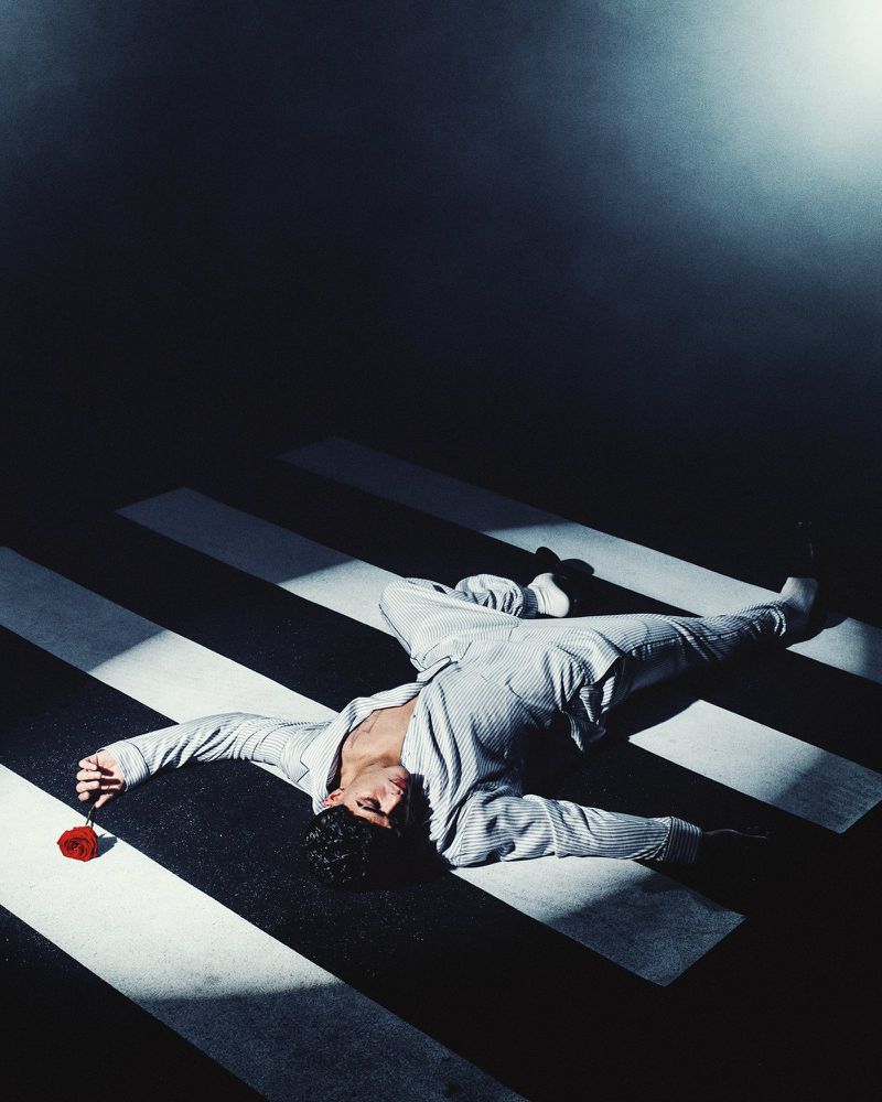A man wearing a grey striped suit lies on a striped crosswalk holding a red rose under dramatic theatrical lighting.