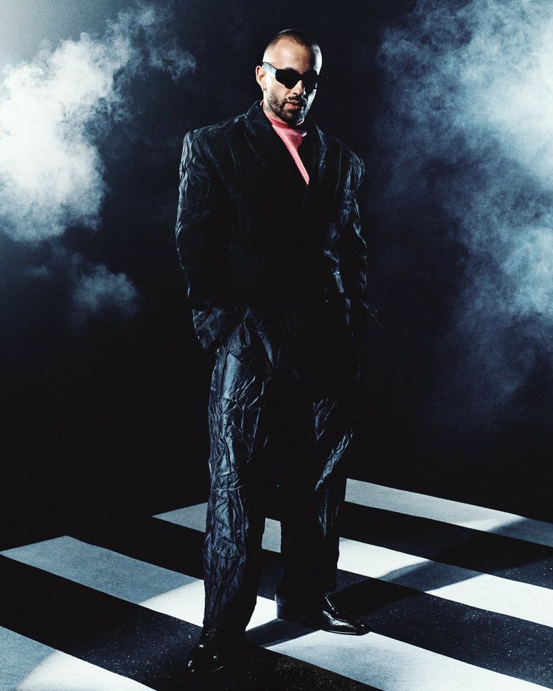 Man wearing dark sunglasses and textured black suit over a pink top against smoky dark background with spotlight bars