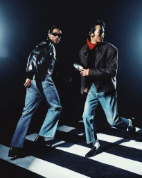 Two male models walking briskly on a striped studio floor wearing dark jackets and blue jeans for Willy Chavarria.