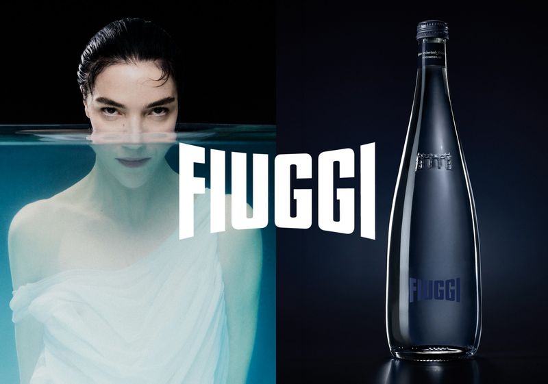 Mariacarla Boscono partially submerged in water beside a glass bottle of Acqua Fiuggi against a dark background