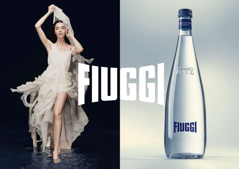 Model Mariacarla Boscono in a flowing white dress standing in water beside a glass bottle of Fiuggi water
