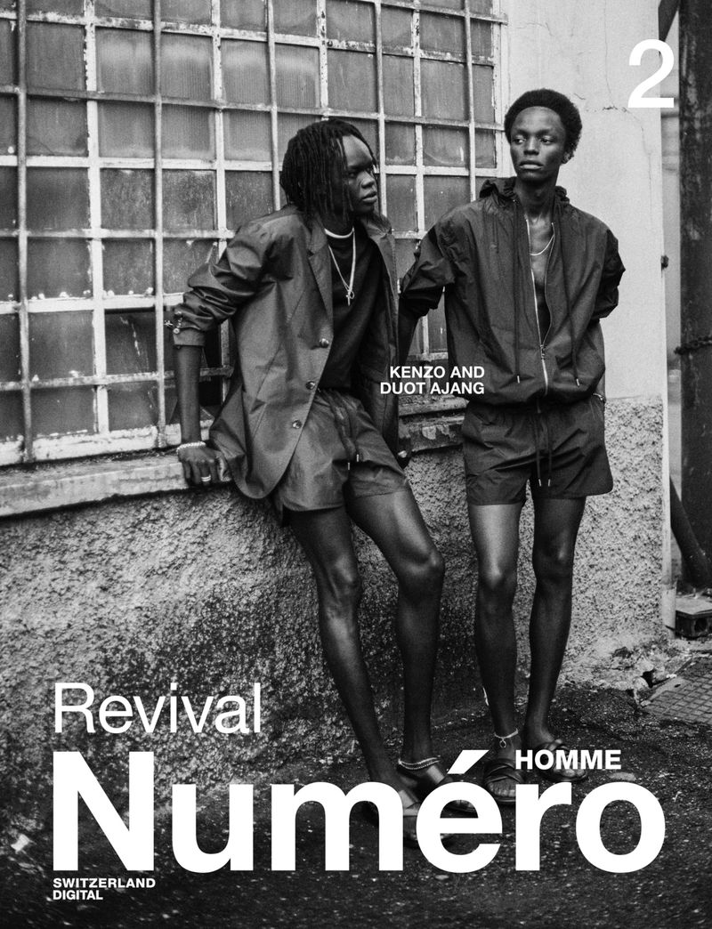 Two dark-skinned men pose in matching outfits in front of a building with a large window; fashion editorial for Numero Homme magazine.