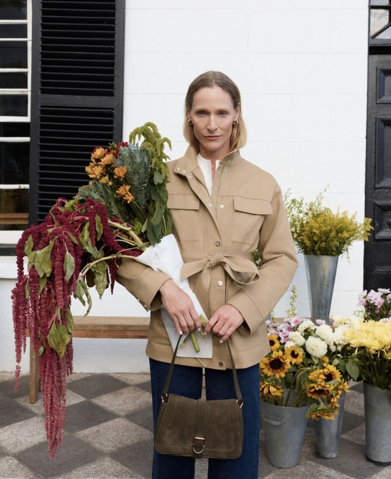 Adult woman posing outdoors with a bouquet of flowers and a brown purse, near a shop with white walls and dark shutters.