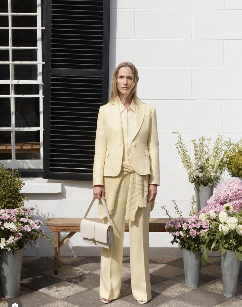 Model wearing a pale yellow suit and holding a white handbag standing against a white building exterior with flowers.