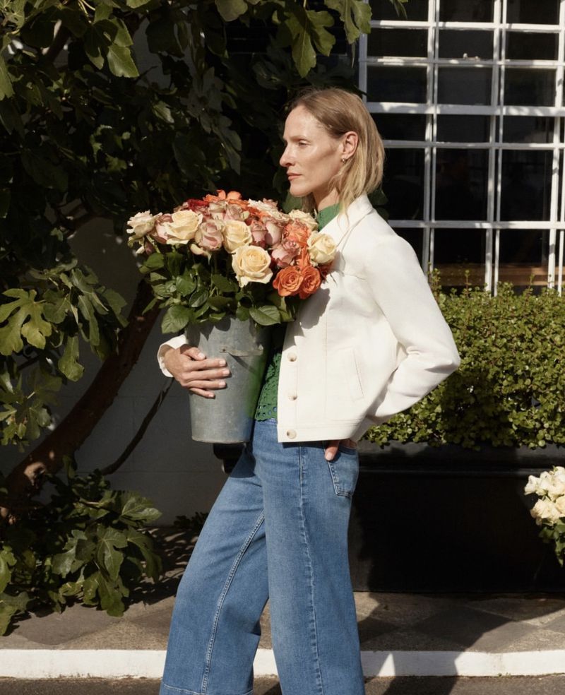 Model carrying a large metal bucket filled with multicolored roses outdoors near a white window frame.