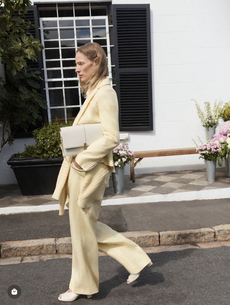 Model wearing a pale yellow suit holding a white handbag walking past a white building exterior