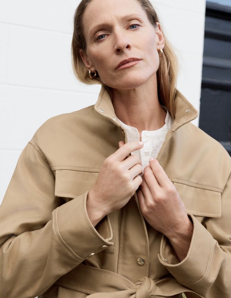 Model wearing a tan jacket and white top adjusting her collar against a white wall