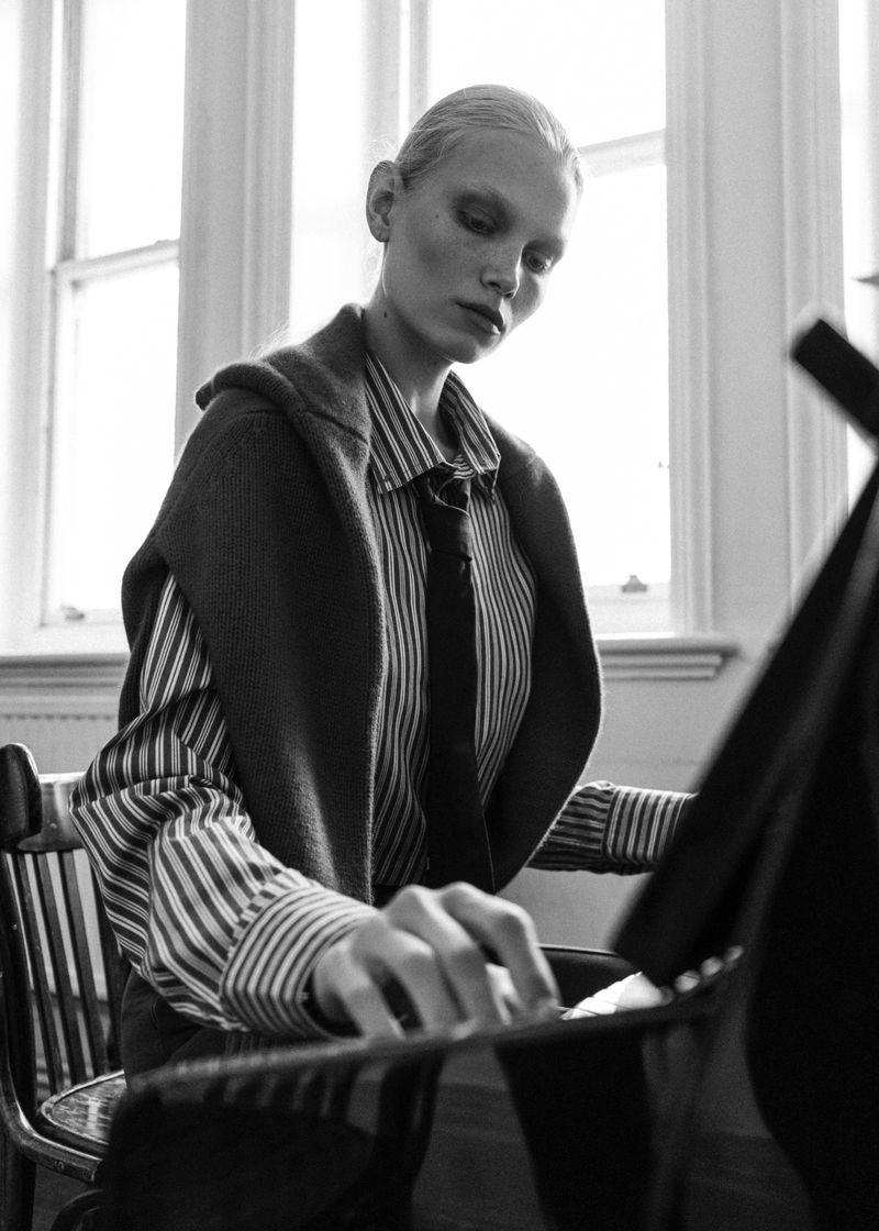 Monochrome portrait of a blonde female model in a striped shirt with a tie, seated at a piano with natural window lighting.