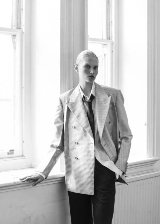 Model with slicked-back hair wearing a double-breasted blazer and dark trousers standing by large windows