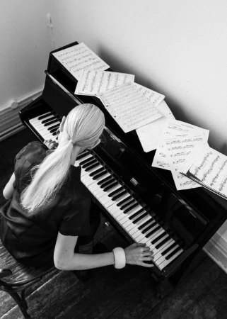 High-angle black and white photograph of a blonde woman playing a piano with sheet music scattered across the instrument.