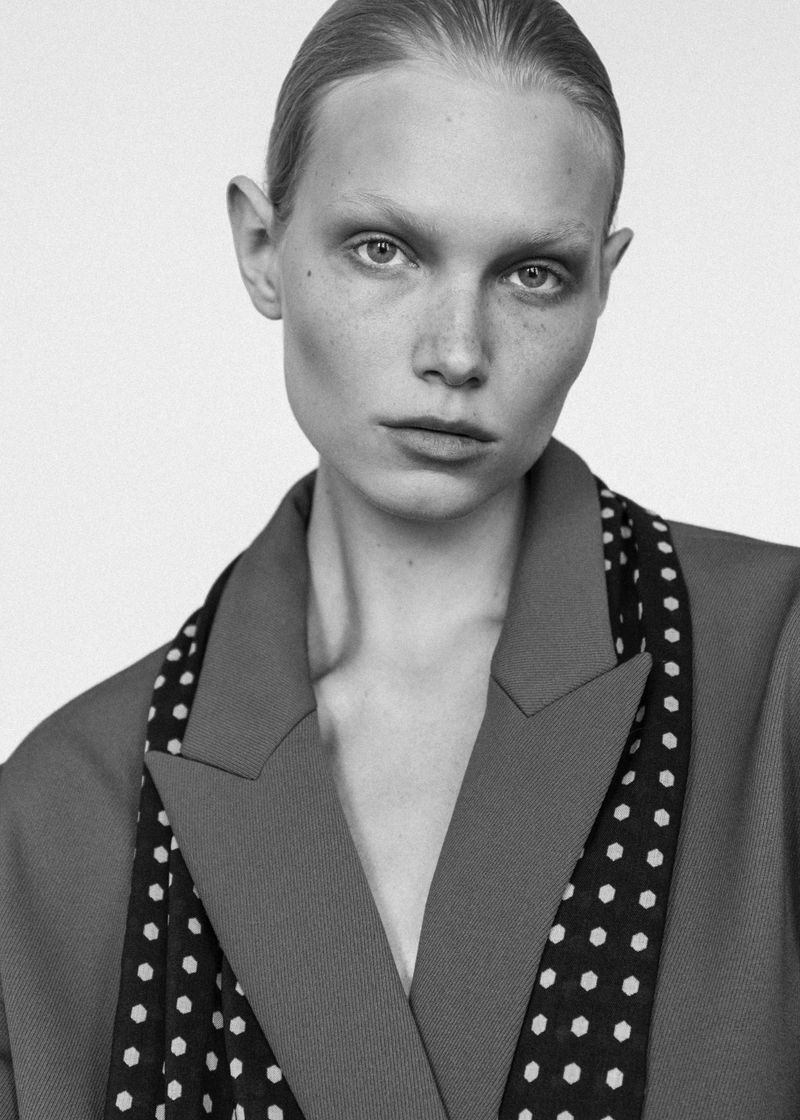 Close-up portrait of a model with sleek hair wearing a tailored jacket and polka dot scarf against a white background
