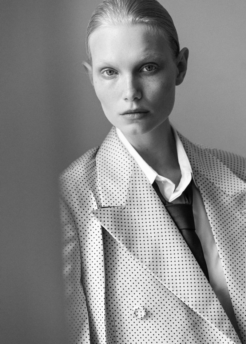 Close-up monochrome portrait of a model wearing a polka dot blazer, white shirt, and tie against a plain studio background.
