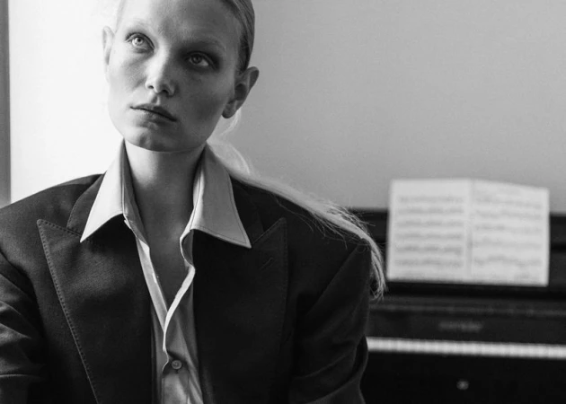 Black and white portrait of a female model wearing a tailored blazer and collared shirt in front of a piano with sheet music.