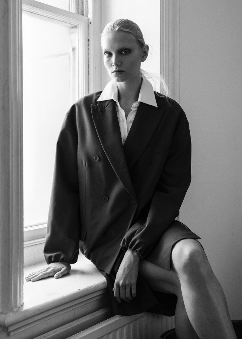 Monochrome portrait of a model sitting on a window sill wearing an oversized blazer and white collared shirt.
