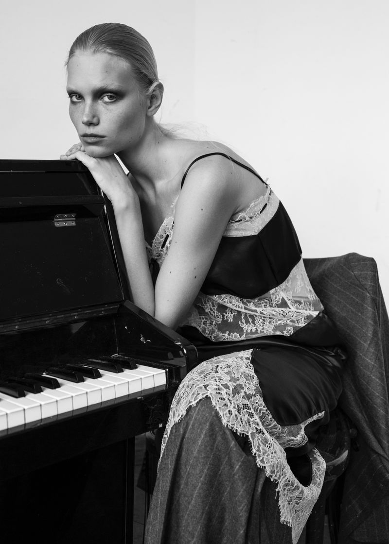 Model leaning on a black upright piano wearing a lace-trimmed slip dress and striped skirt against a plain wall