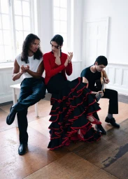 Three models posing in a bright room with hardwood floors, featuring a woman in a ruffled black and red skirt clapping beside two seated men.