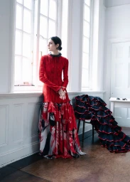 Model standing by a tall window wearing a red textured dress with a patterned skirt in an interior room.