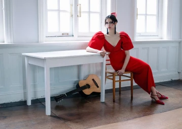 Model in vibrant red dress seated on a wooden chair next to a white table with a classical guitar underneath.