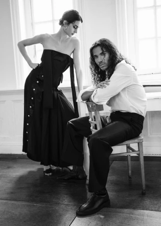 Black and white editorial portrait of a woman standing and a man seated on a chair indoors near large windows.