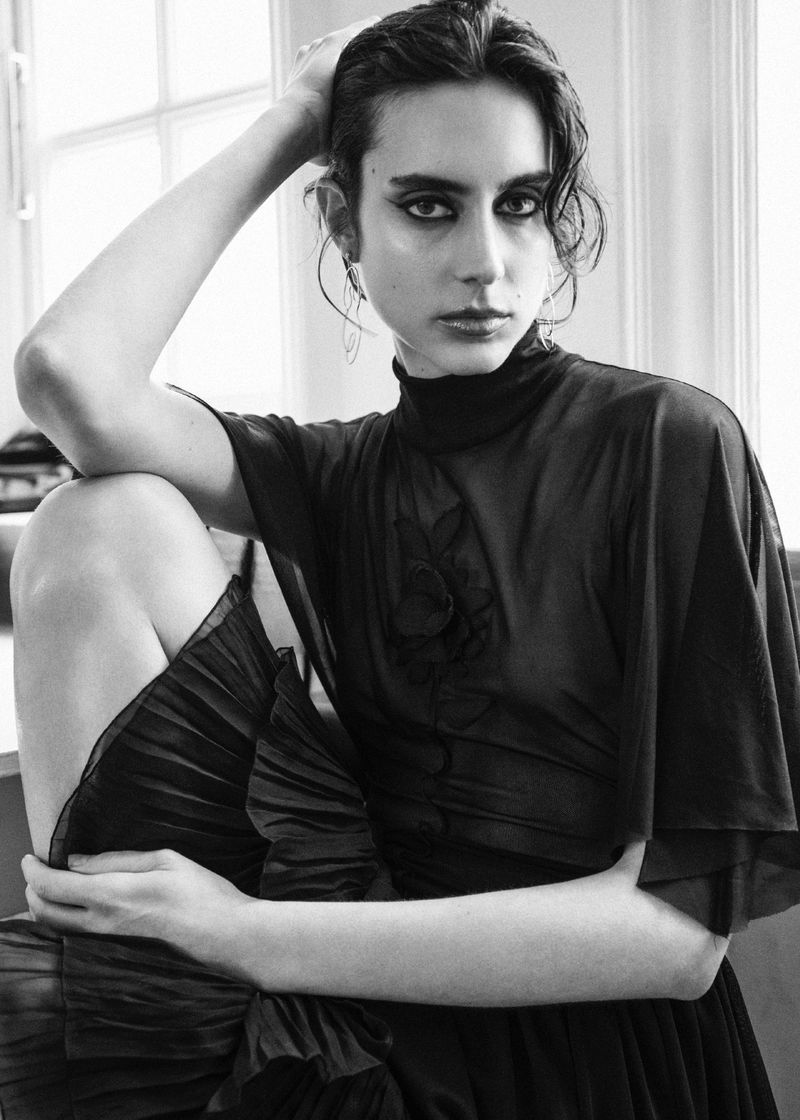 Monochrome portrait of a model in a dark sheer mock-neck top with pleated skirt against a window.