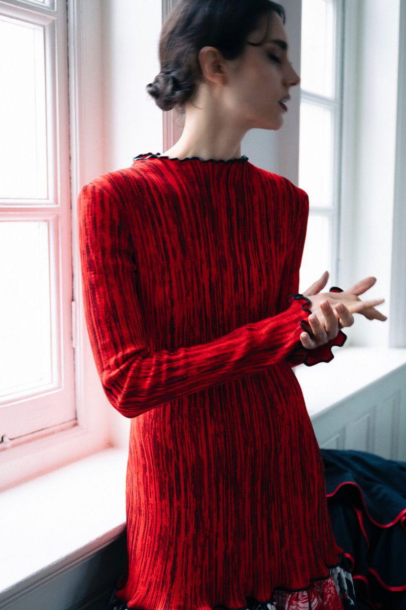 Model in a textured red long-sleeve dress posing near large white windows for 5ELEVEN Magazine