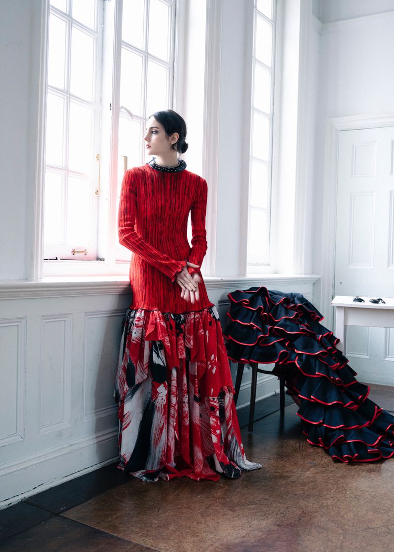 Model standing by a tall window wearing a red textured dress with a patterned skirt in an interior room.
