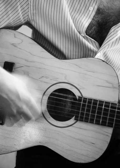 Monochrome close-up of a musician playing an acoustic guitar with a white soundboard and dark fretboard, wearing a striped shirt.