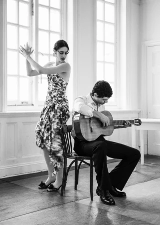 Black and white editorial image of a woman dancing while a man sits playing an acoustic guitar indoors.