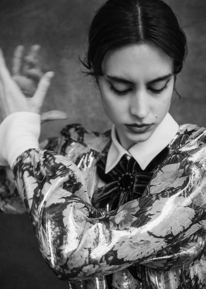 Black and white portrait of a young woman in a floral top, looking down with her hands raised in an elegant pose against a dark background.