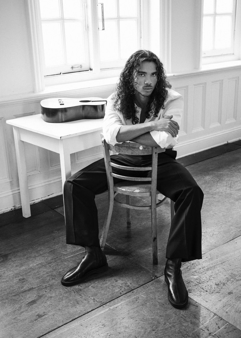 Man with long curly hair seated backwards on a wooden chair, wearing a white shirt and dark trousers in an interior room.