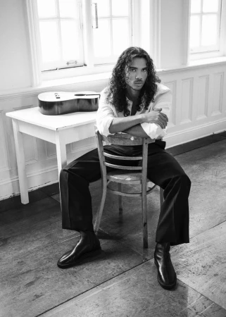 Man with long curly hair seated backwards on a wooden chair, wearing a white shirt and dark trousers in an interior room.