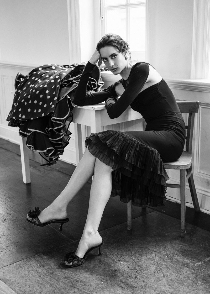 Monochrome fashion editorial featuring a seated model in a black dress next to a table with a polka-dot dress.