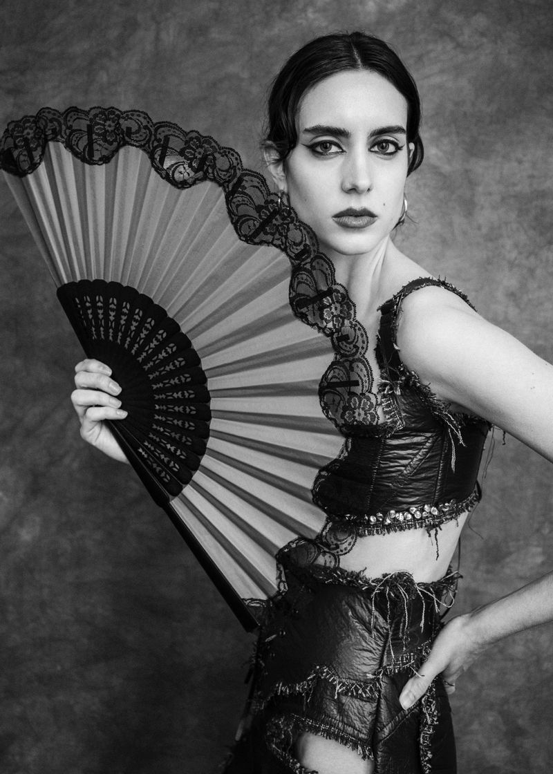 Model holding a black lace folding hand fan wearing a dark textured top against a mottled grey background