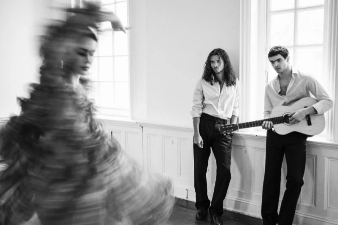 Monochrome fashion editorial featuring a blurred dancer in motion and two men by a window with a guitar