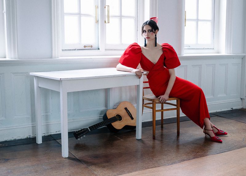 Model in vibrant red dress seated on a wooden chair next to a white table with a classical guitar underneath.