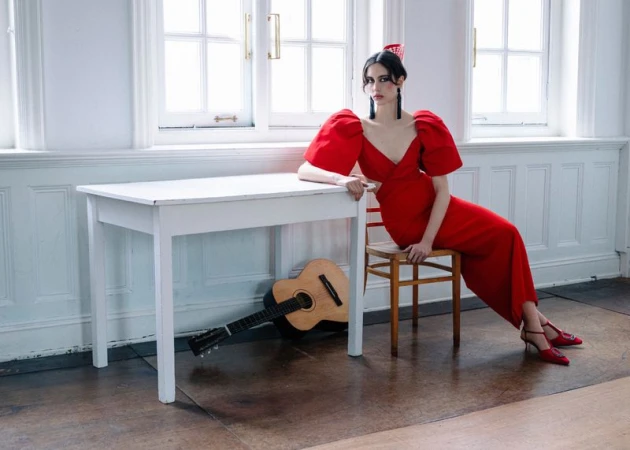 Model in vibrant red dress seated on a wooden chair next to a white table with a classical guitar underneath.