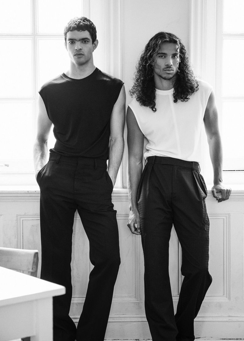 Two male models posing indoors by a window in contrasting black and white sleeveless tops and dark trousers for 5ELEVEN Magazine.