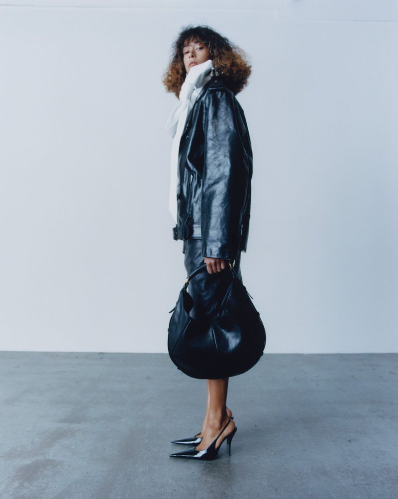 Model in a black leather jacket and matching skirt holding a black handbag against a light grey studio background.