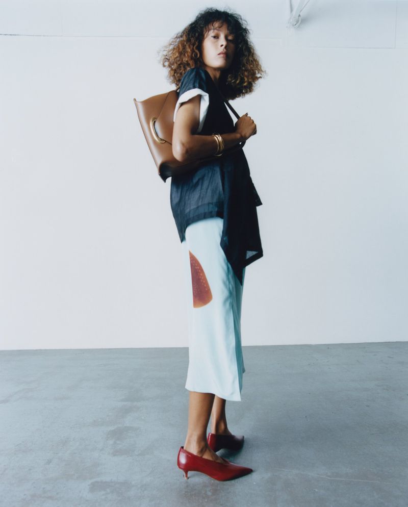 Model with curly hair wearing a dark top, light skirt, and red shoes while carrying a brown leather bag