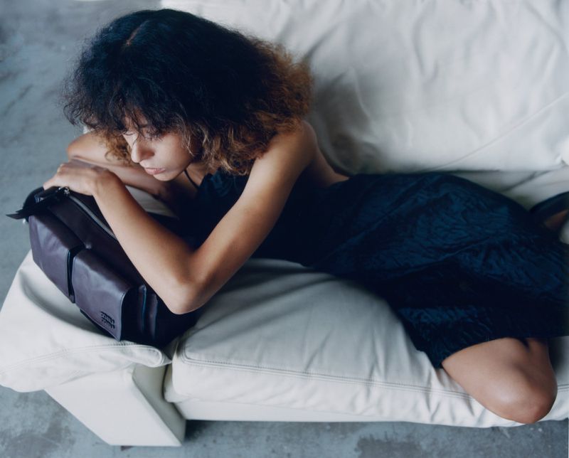 Model lying on a white sofa holding a dark leather bag in an editorial portrait for Puss Puss Magazine by Maya Skelton