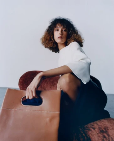 Model with curly hair crouching on a reddish-brown sofa holding a large tan leather handbag against a plain background